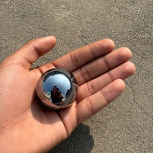 Puede incluir: Una esfera pulida, similar al cromo, que refleja el entorno. El objeto esférico se sostiene en una mano abierta, mostrando su superficie lisa y reflectante. La superficie de la esfera refleja el cielo y la persona que la sostiene.