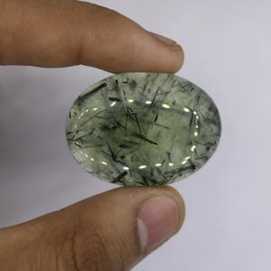 Könnte beinhalten: Ovaler Edelstein mit transluzentem, hellgrünem Farbton. Der Stein weist dunkle, nadelförmige Einschlüsse auf, die ein einzigartiges Muster bilden. Auf weißem Hintergrund zwischen Fingern gehalten, reflektiert die polierte Oberfläche das Licht.