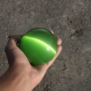 Puede incluir: Una esfera verde vibrante con un efecto ojo de gato luminoso. La gema pulida y redonda se sostiene en una mano, mostrando su superficie lisa y su brillo interno. El color de la esfera es un verde brillante y saturado.