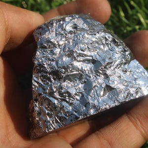 May include: A piece of raw, metallic-looking material with a reflective, textured surface. The object is held in a hand, with the green grass in the background.