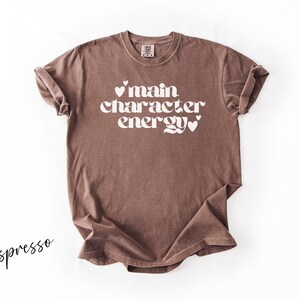 May include: Espresso-colored t-shirt with the phrase "main character energy" in white, stylized font. The text includes heart symbols. The shirt has a crew neck and short sleeves.