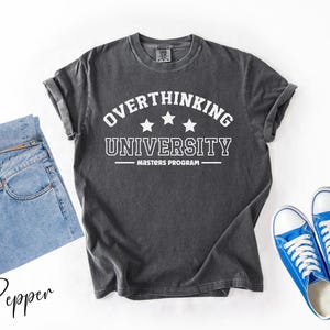 May include: Dark gray t-shirt with the text "OVERTHINKING UNIVERSITY MASTERS PROGRAM" in white. The shirt is paired with light blue jeans and blue sneakers with white laces and soles. The items are arranged on a white surface.