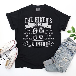 May include: Black t-shirt with white text that reads "THE HIKER'S CODE: TAKE NOTHING BUT PICTURES, LEAVE NOTHING BUT FOOTPRINTS, KILL NOTHING BUT TIME." The shirt is paired with white sneakers and blue jeans.