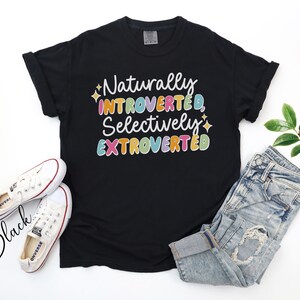 May include: Black t-shirt with the text "Naturally Introverted, Selectively Extroverted" in colorful bubble letters. The shirt is paired with light-wash denim jeans and white canvas sneakers.