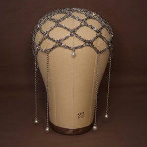 May include: A silver headpiece with a chainmail design, decorated with small sequins and dangling chains. Each chain ends with a faux pearl. The headpiece is displayed on a mannequin head.