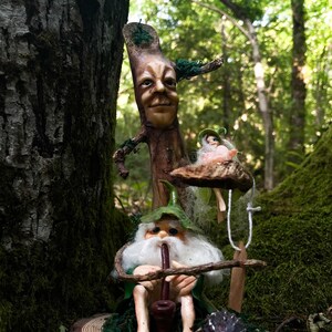 Può includere: Una scena boschiva fantastica con un albero di legno scolpito con un volto, uno gnomo che fuma la pipa e una piccola fata. Lo gnomo indossa un cappello e una veste verdi. La scena è ambientata su un tronco muschioso con un piccolo riccio.