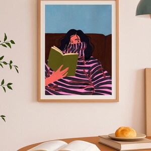 Peut inclure: Impression d'art encadr&eacute;e d'une personne lisant un livre. L'illustration repr&eacute;sente une personne aux cheveux noirs portant un pull &agrave; rayures roses et noires. Le fond est bleu et marron. Une table en bois avec un livre ouvert et une assiette avec un petit pain au premier plan.