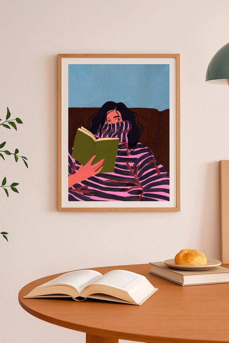 Peut inclure: Impression d'art encadr&eacute;e d'une personne lisant un livre. L'illustration repr&eacute;sente une personne aux cheveux noirs portant un pull &agrave; rayures roses et noires. Le fond est bleu et marron. Une table en bois avec un livre ouvert et une assiette avec un petit pain au premier plan.