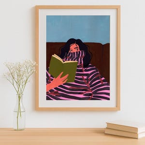 Peut inclure: Une impression d'art encadr&eacute;e repr&eacute;sentant une personne lisant un livre. La personne porte un pull &agrave; rayures roses et noires. L'arri&egrave;re-plan pr&eacute;sente un canap&eacute; marron et un ciel bleu. L'impression d'art est dans un cadre en bois brun clair.