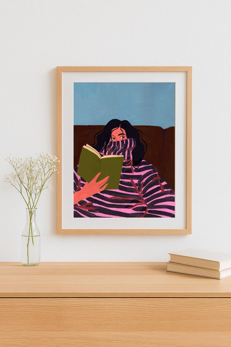 Peut inclure: Une impression d'art encadr&eacute;e repr&eacute;sentant une personne lisant un livre. La personne porte un pull &agrave; rayures roses et noires. L'arri&egrave;re-plan pr&eacute;sente un canap&eacute; marron et un ciel bleu. L'impression d'art est dans un cadre en bois brun clair.