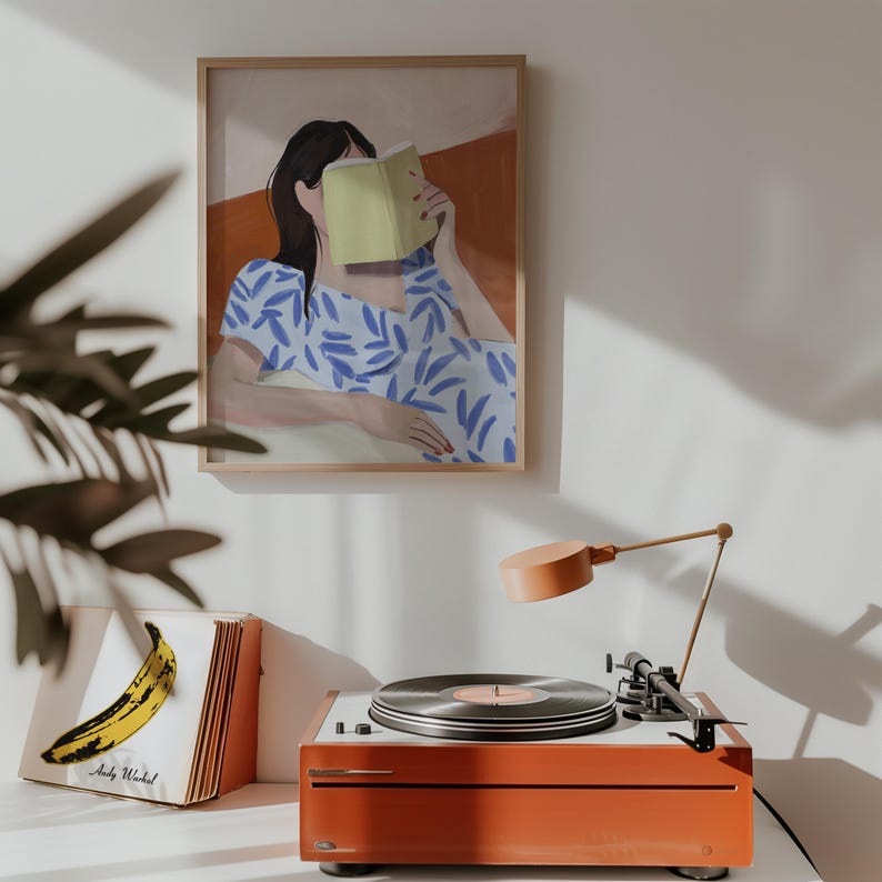 May include: A framed art print depicts a person reading a book, with a blue and white patterned dress. A vintage orange record player with a vinyl record is on a white surface. A lamp and a record album with a banana graphic are also present.