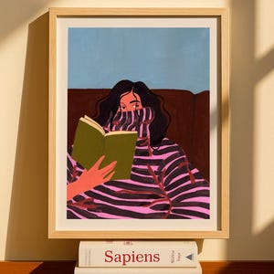Peut inclure: Impression d'art encadr&eacute;e d'une personne lisant un livre, v&ecirc;tue d'un pull &agrave; rayures roses et noires. L'&oelig;uvre est dans un cadre en bois clair. Trois livres sont empil&eacute;s en dessous, avec les titres "Sapiens", "Before We Were Yours" et "Educated".