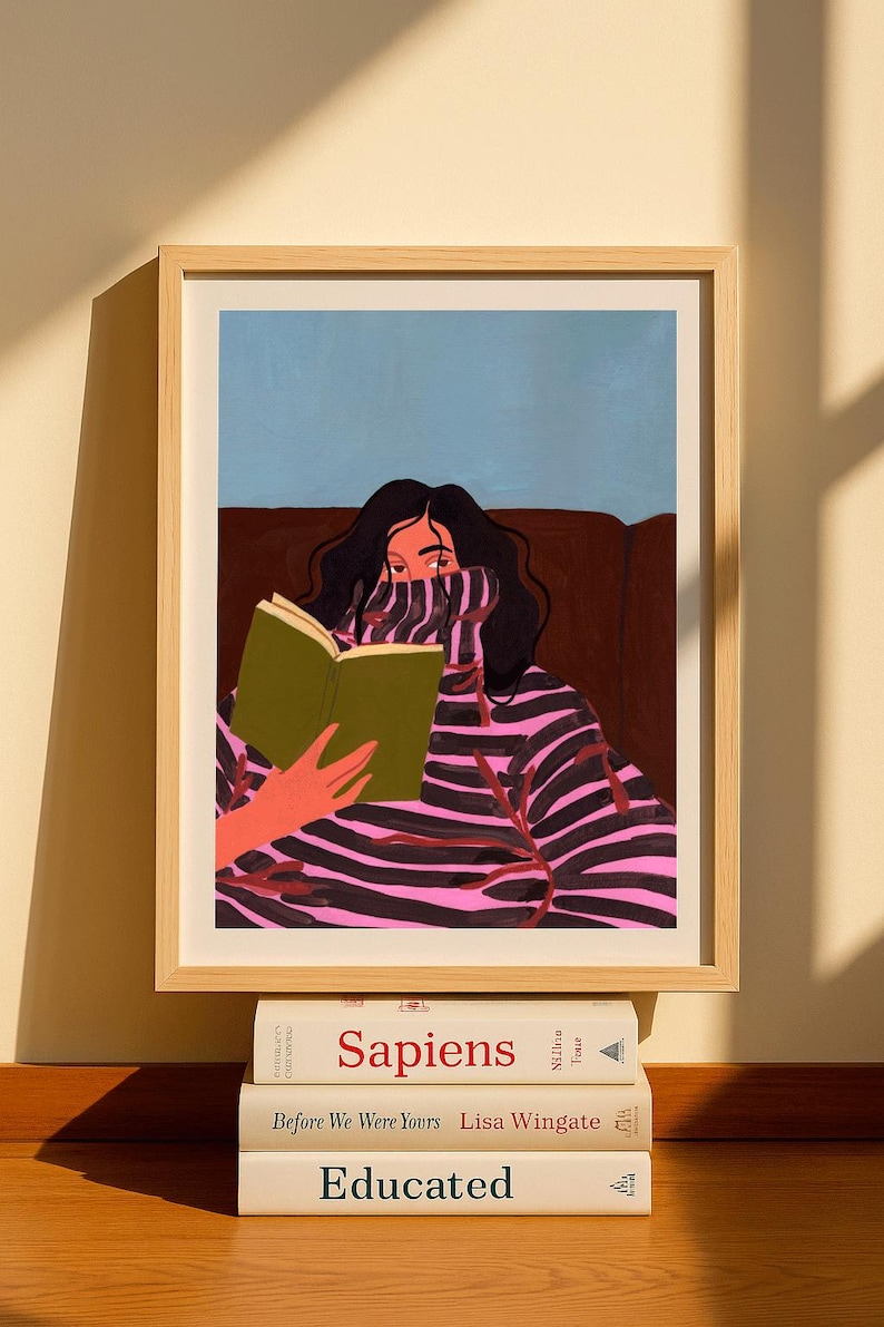 Peut inclure: Impression d'art encadr&eacute;e d'une personne lisant un livre, v&ecirc;tue d'un pull &agrave; rayures roses et noires. L'&oelig;uvre est dans un cadre en bois clair. Trois livres sont empil&eacute;s en dessous, avec les titres "Sapiens", "Before We Were Yours" et "Educated".