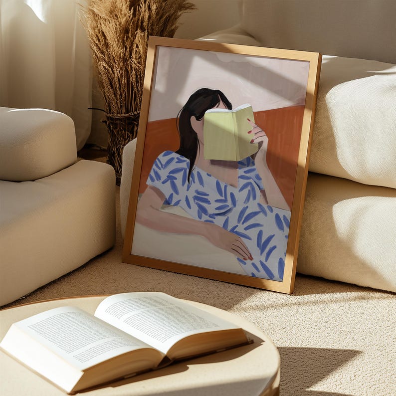 May include: A framed art print depicts a person reading a book. The artwork features a woman in a blue and white patterned dress, with a green book covering her face. The frame is a light wood color.
