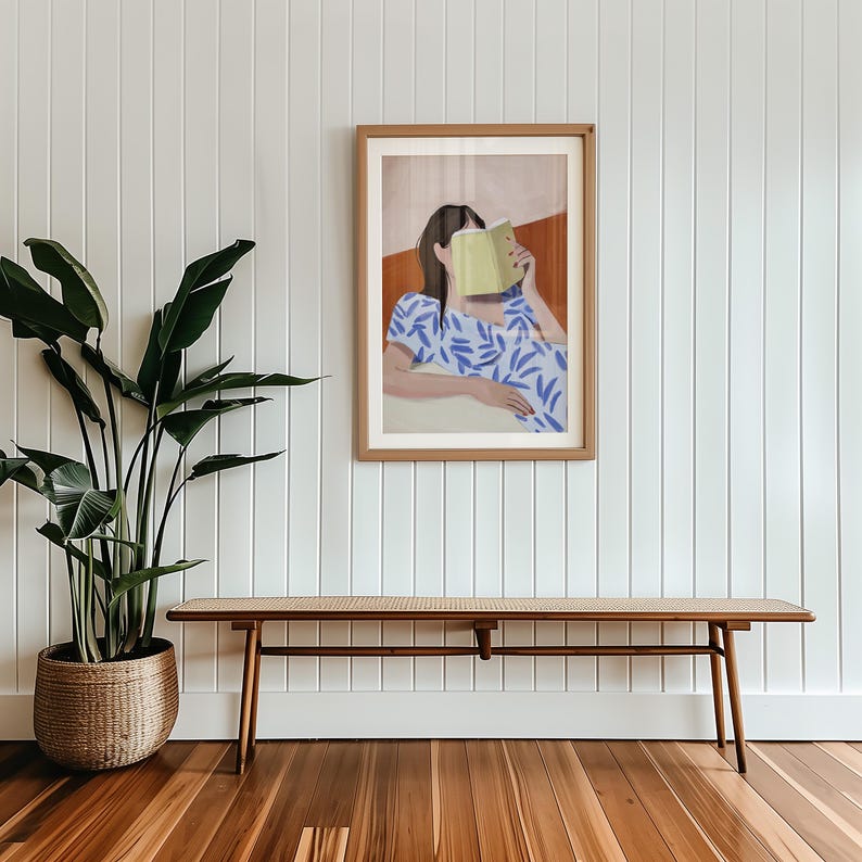 May include: A framed art print depicts a person reading a book, with the book obscuring their face. The person is wearing a blue and white patterned dress. The artwork hangs on a white wall above a wooden bench with a woven top. A large potted plant is to the left.