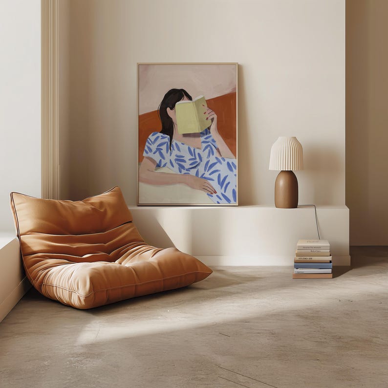 May include: An orange, low-profile, tufted lounge chair sits on a concrete floor. A framed artwork depicts a person reading a book, with a blue and white patterned dress. A brown table lamp and a stack of books are also visible.