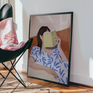 May include: A framed art print depicts a person reading a book, with the book obscuring their face. The person is wearing a blue and white patterned dress. The background features warm tones of orange and beige.