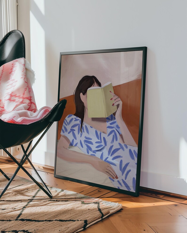 May include: A framed art print depicts a person reading a book, with the book obscuring their face. The person is wearing a blue and white patterned dress. The background features warm tones of orange and beige.