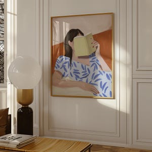 May include: An art print depicts a person reading a book, with a blue and white patterned dress. The artwork is framed in gold and hangs on a white wall. A modern lamp with a white globe and a black base is in the foreground.