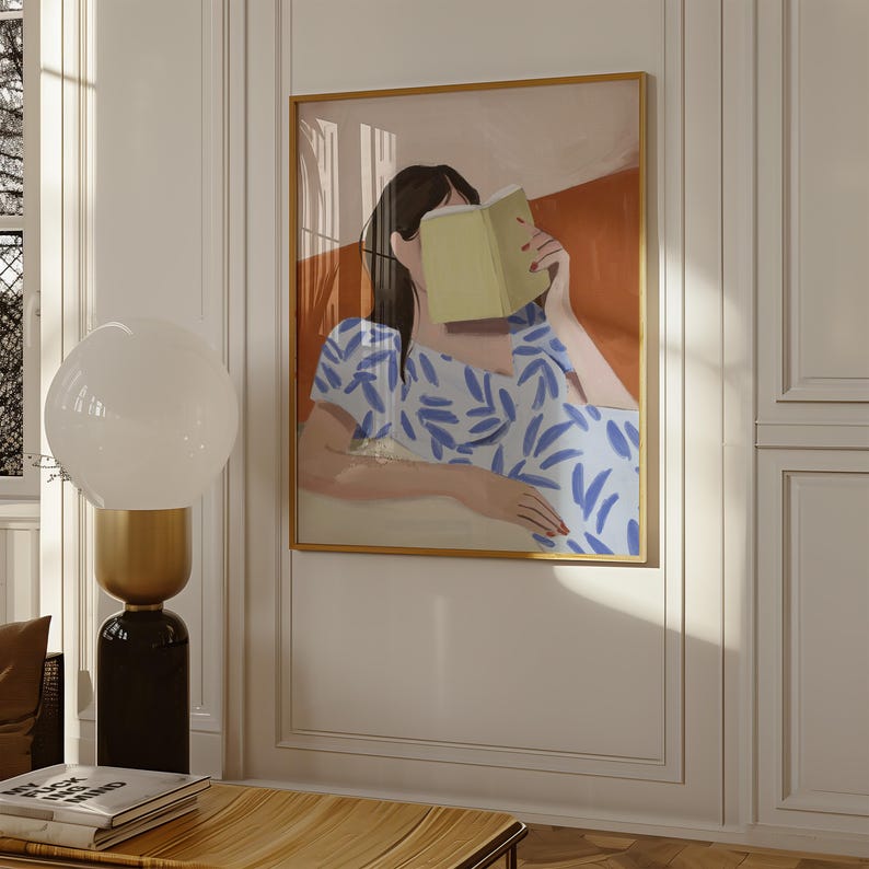 May include: An art print depicts a person reading a book, with a blue and white patterned dress. The artwork is framed in gold and hangs on a white wall. A modern lamp with a white globe and a black base is in the foreground.