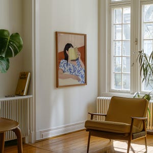 May include: A framed art print depicts a person reading a book, with the book obscuring their face. The person is wearing a blue and white patterned top. The artwork hangs on a white wall in a room with a wooden floor and a mustard-colored armchair.