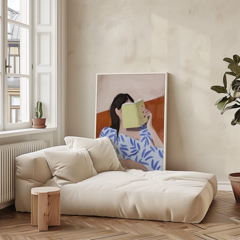 May include: A cream-colored, low-profile sofa with two matching pillows and a small wooden side table. A framed art print depicts a person reading a book, with a blue and white patterned dress. A potted plant is in the background.