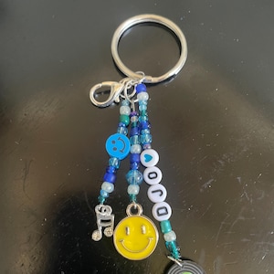 May include: A silver keyring with a beaded charm. The charm features a yellow smiley face, a miniature vinyl record, and the word "DOJO" spelled out with white and teal beads. Additional charms include a musical note and blue smiley face beads.