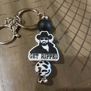 Pode incluir: Um chaveiro com um anel prateado e um design de contas em preto e branco. O chaveiro apresenta um gráfico em preto e branco de um homem usando um chapéu de cowboy e óculos de sol, com o texto "GET RIPPED".