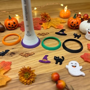 Könnte beinhalten: Ein weißer Philips Sonicare elektrische Zahnbürstengriff mit einem violetten Basisring, begleitet von drei zusätzlichen austauschbaren Basisringen in Orange, Grün und Schwarz, umgeben von verschiedenen kleinen Halloween-Dekorationen.