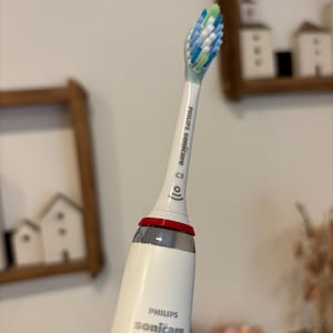 Könnte beinhalten: Eine weiße elektrische Zahnbürste von Philips Sonicare mit einem roten Akzentring und einem Bürstenkopf mit blauen, grünen und weißen Borsten. Der Markenname ist auf dem Griff aufgedruckt.