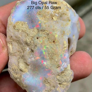 May include: A raw opal stone with a rough, textured exterior. The opal displays iridescent colors, including red, green, and blue, within its milky white surface. The text "Big Opal Raw 277 cts / 55 Gram" is visible.