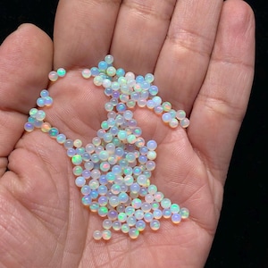 May include: A handful of small, round opals. The opals are mostly white with iridescent flashes of blue, green, and yellow. The gemstones are smooth and polished, and appear to be of high quality. The opals are scattered across the palm of a hand.