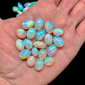 Może przedstawiać: Kolekcja owalnych opali o opalizujących barwach, takich jak niebieski, zielony i żółty. Opale są trzymane w otwartej dłoni na czarnym tle. Kamienie szlachetne różnią się rozmiarem i wykazują grę barw.