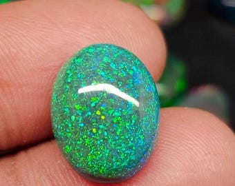 May include: An oval gemstone, predominantly green with shimmering blue and yellow inclusions. The polished surface catches the light, creating a sparkling effect. The gem is held between fingers.