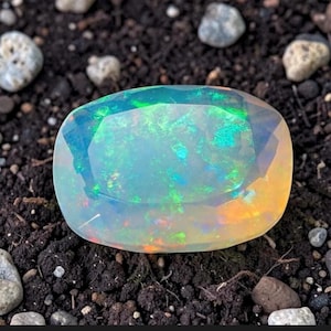 Puede incluir: Una gran piedra preciosa de ópalo de forma ovalada, con una apariencia translúcida e iridiscente. La piedra muestra un juego de colores con destellos de tonos verdes, azules y anaranjados. El ópalo se encuentra sobre un fondo de tierra oscura y pequeñas rocas.
