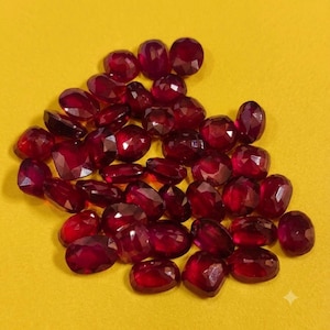 Puede incluir: Una colección de numerosos rubíes sueltos, facetados, de color rojo intenso y talla brillante. Cada piedra preciosa muestra una calidad translúcida y una superficie pulida, dispuestas en un grupo sobre un fondo amarillo brillante y sólido.