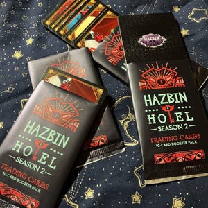 以下が含まれることがあります： Hazbin Hotel Season 2の黒いトレーディングカードブースターパックが複数あり、それぞれ10枚のカードが入っています。パッケージには赤とライトグリーンのグラフィックデザインが施されており、目のアイコンと「Hazbin Hotel Season 2 Trading Cards 10-Card Booster Pack」という文字が記載されています。