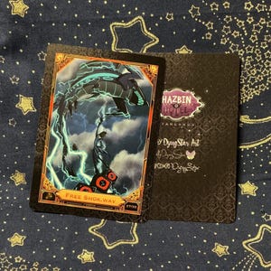 May include: A rectangular trading card featuring a dark illustration of a creature with lightning, labeled Free Shok.wav, alongside a black card back with the Hazbin Hotel logo and text by Dying Star Art, both with ornate gold and black borders.