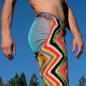 May include: A person wearing denim pants with a colorful, zig-zag crochet pattern on the right leg. The crochet design features a vibrant mix of red, orange, yellow, green, blue, and black. The person is wearing brown and tan patterned boots.