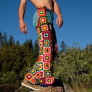 May include: Colorful crochet leg warmers with a patchwork design. The leg warmers feature squares of red, green, yellow, blue, and white. The person is wearing blue jeans and brown shoes. The background is a clear blue sky and trees.