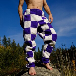 May include: Hand-crocheted pants in a checkerboard pattern of white and purple squares. The pants are loose-fitting with a drawstring at the ankles. The background features a blue sky and trees.