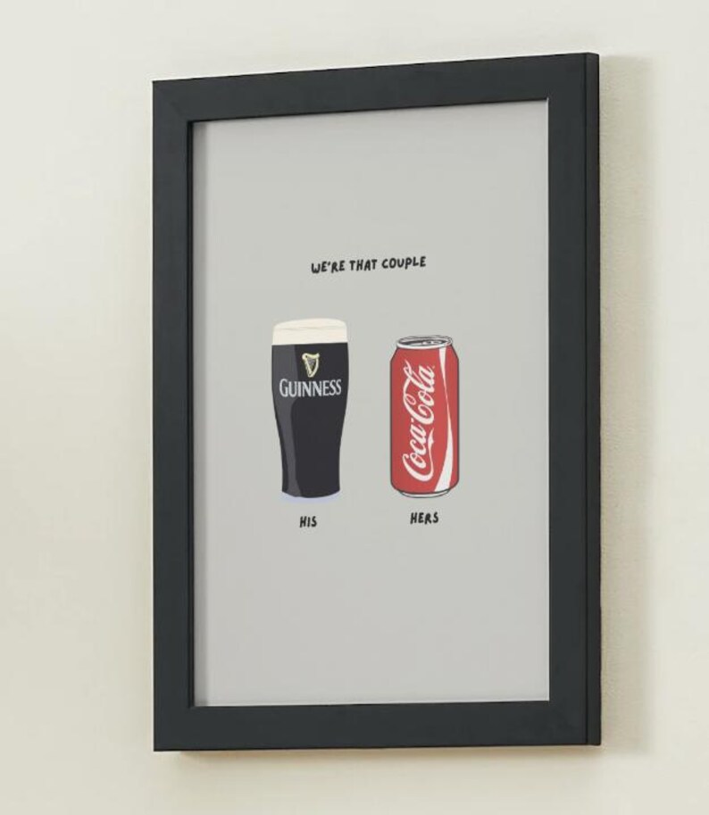Puede incluir: Impresi&oacute;n enmarcada con fondo gris. Presenta un vaso de pinta Guinness etiquetado "HIS" y una lata de Coca-Cola etiquetada "HERS". El texto "WE'RE THAT COUPLE" est&aacute; encima de las im&aacute;genes. El marco es negro.