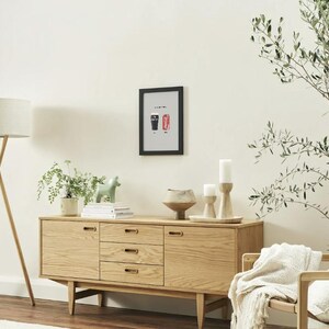 Puede incluir: Un aparador de madera clara con tres cajones y dos puertas, coronado con una impresi&oacute;n enmarcada, velas y un cuenco de cer&aacute;mica. Una l&aacute;mpara de pie con una pantalla beige est&aacute; a la izquierda.