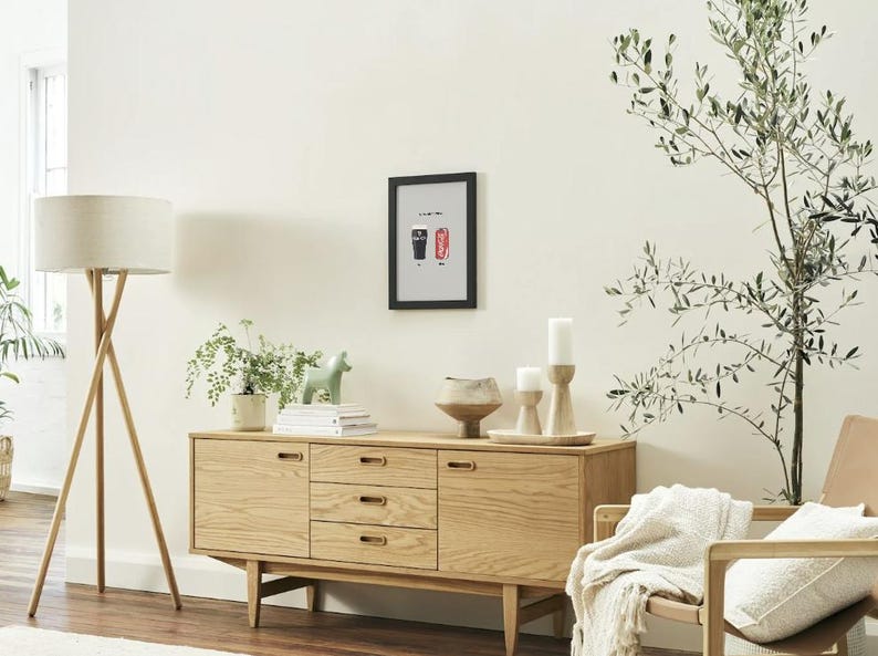 Puede incluir: Un aparador de madera clara con tres cajones y dos puertas, coronado con una impresi&oacute;n enmarcada, velas y un cuenco de cer&aacute;mica. Una l&aacute;mpara de pie con una pantalla beige est&aacute; a la izquierda.