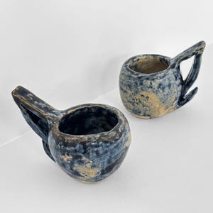 May include: Two handmade ceramic cups with a unique design. The cups are a mix of blue and tan, with a textured surface. Each cup has a handle and a spout-like extension. The cups are placed on a white surface.