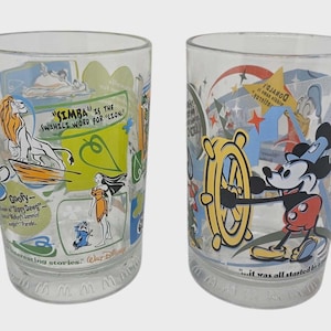 May include: Two clear drinking glasses with colorful Disney character designs. One glass features Simba, Goofy, and Moana. The other glass has Mickey Mouse holding a ship's wheel. Text includes "SIMBA" and "it was all started".