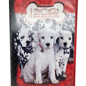 Puede incluir: Una carátula de DVD de la película "102 Dálmatas" de Disney. La portada presenta tres cachorros dálmatas en primer plano, con un fondo de otros dálmatas. El título se muestra en un diseño en forma de hueso.