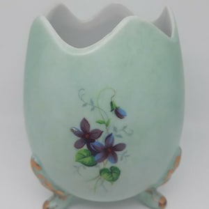 May include: A light green, egg-shaped ceramic vase with a scalloped rim and three small feet. The vase is decorated with a floral design featuring purple flowers and green leaves. The feet have gold accents.
