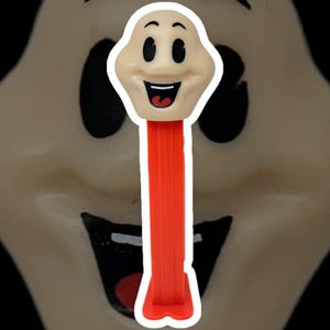 May include: A PEZ dispenser with a smiling cartoon face, light beige in colour, and a bright red dispenser. The face has black eyes and an open mouth. The background is a blurred image of a similar face.