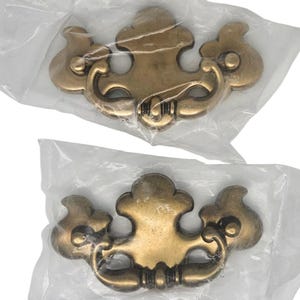 May include: Two brass-toned metal drawer pulls with a decorative, ornate shape. Each pull features a central mounting plate with a curved, swinging handle attached. The hardware is individually packaged in clear, crinkled plastic protective bags.